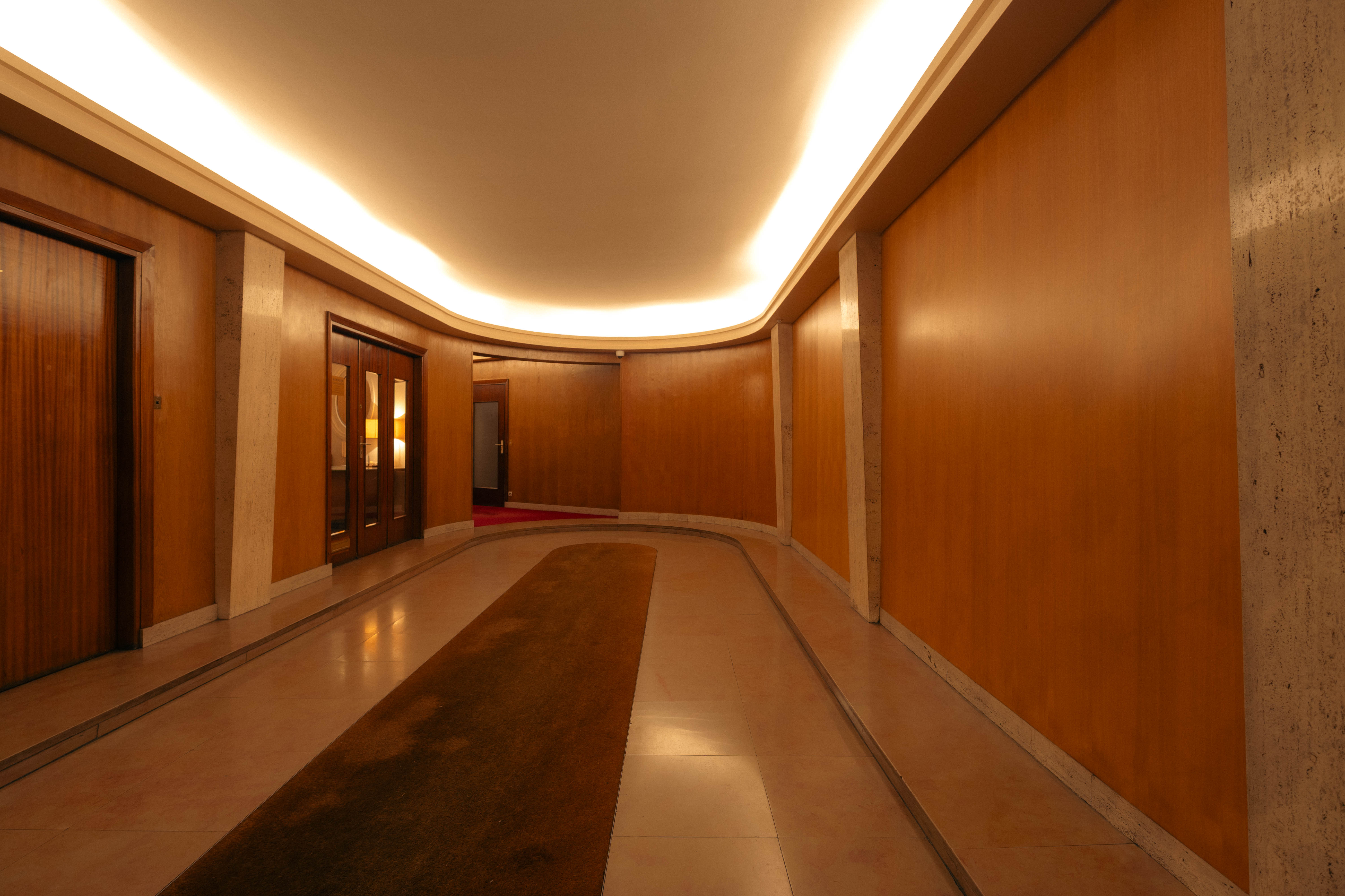Couloir courbe grand bâtiment, murs teck et travertin, éclairage indirect
