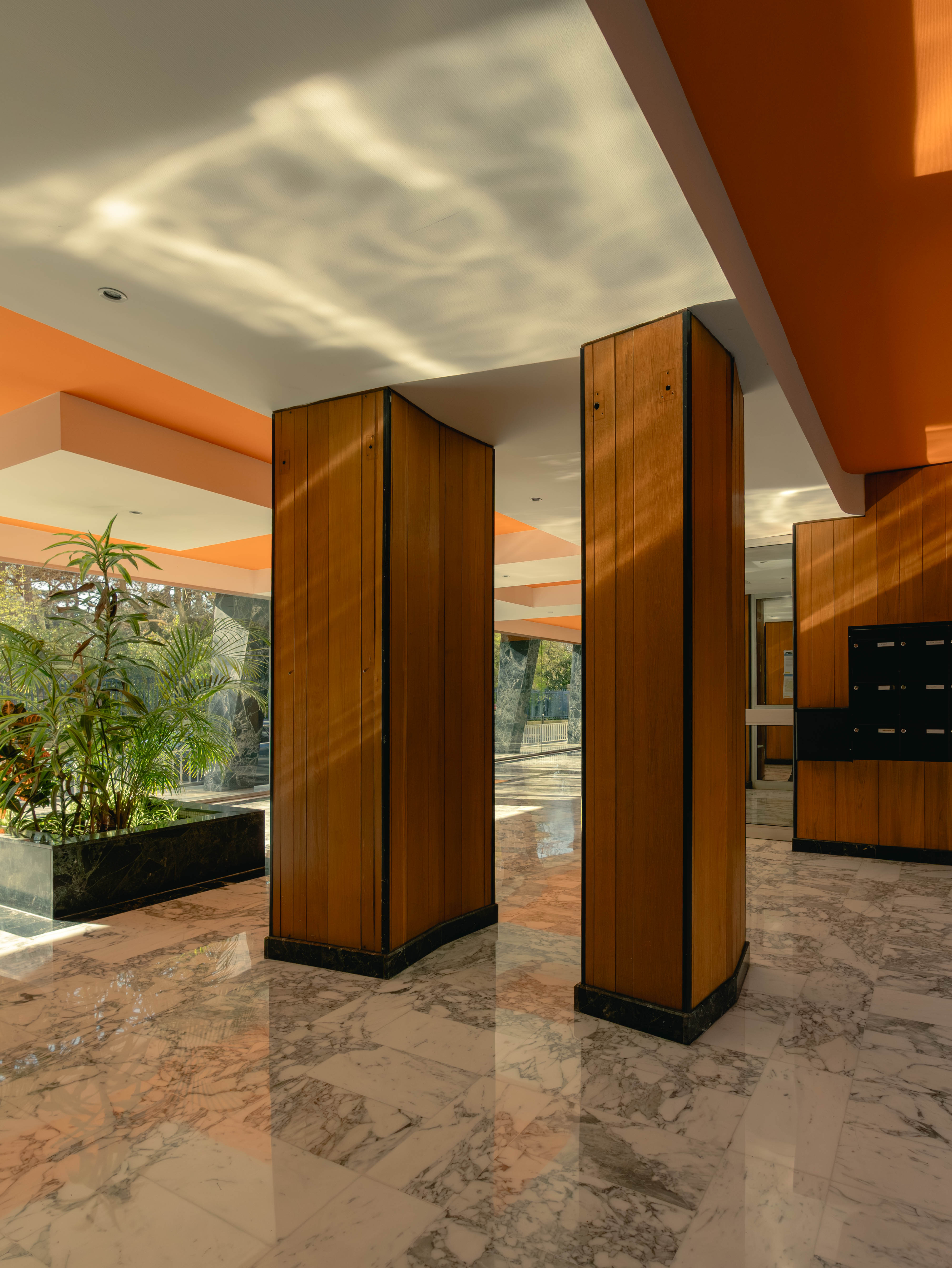 Hall colonnes bois, plafond orange, sol marbre rose, végétaux tropicaux
